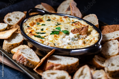 Close up view of an artichoke dip surrounded by various breads and crackers for dipping.