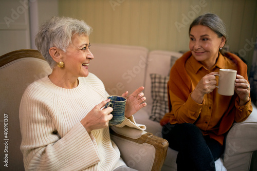 Stylish female friends on retirement sitting comfortably indoors with large mugs, drinking coffee, discussing future plans or recollecting good old days, enjoying nice time together, smiling