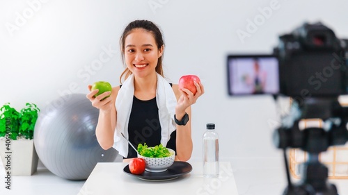 Beautiful asian woman healthy blogger is showing apple fruit and clean diet food. In front of the camera to recording vlog video live streaming at home.Fitness influencer on social media online.