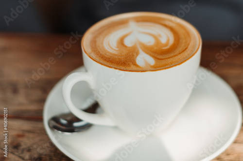 Closeup shot of white cup filled with aromatic coffee with heart shape latte art foam on wood table near window with light shade on tabletop at cafe with spoon besides it