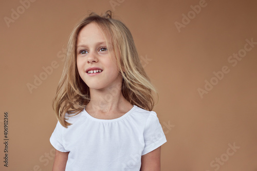 Closeup front portrait of adorable little girl with wavy blond hair, dressed in white clothes, over beige background.