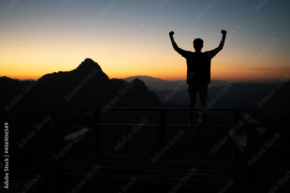 Silhouette of young Asian man standing on a bench with raising fists to the sky as a winner while admires the fantastic landscape of mountain peaks with a soft golden horizon line on sunset or sunrise