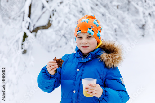 Adorable kid boy drinking hot chocolate and eating brownie cake in winter forest. Happy healthy child with cup of steaming cocoa or tea, outdoors. Active family leisure, walking in snow landscape photo