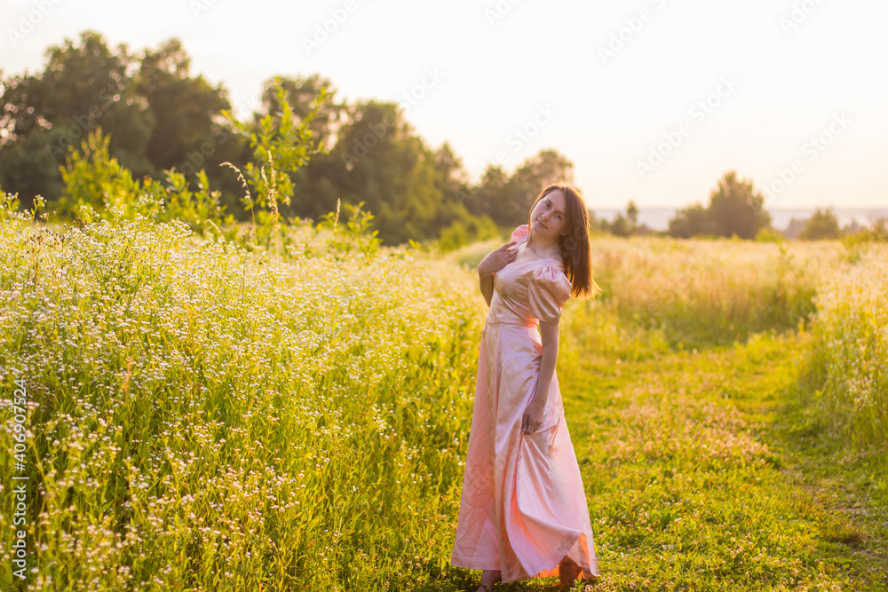 Fototapeta premium Beautiful photo of woman with dark hair standing in the wildflowers field barefoot holding the bottom of the long pink dress on a warm summer evening. Joyfull summer walking. 