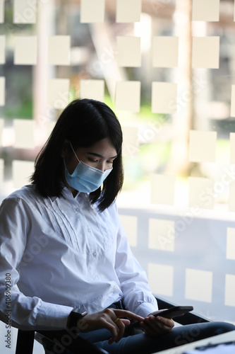 Business woman wearing protective mask taking a break at her workspace and using mobile phone.