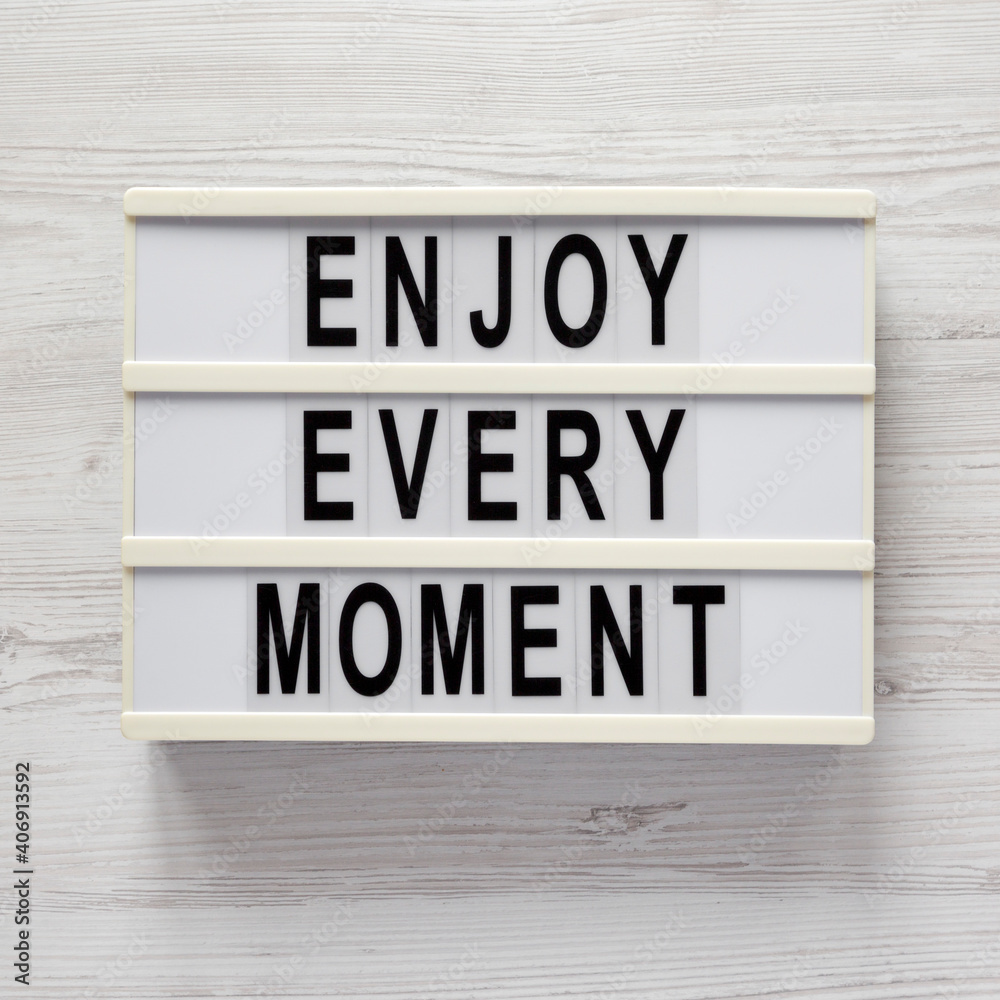 'Enjoy every moment' on a lightbox on a white wooden surface, top view. Flat lay, overhead, from above. 'Enjoy every moment' on a lightbox on a white wooden surface, top view. Flat lay, overhead, from above.