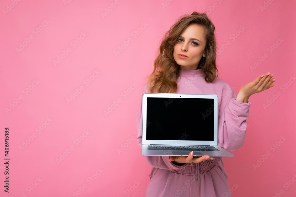 Close-up portrait of beautiful young dark blond curly hair woman holding computer laptop with empty copy space looking at camera and thinking about something having doubts posing hand isolated on pinl
