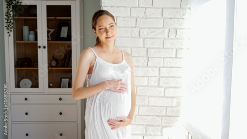 Smiling pregnant lady in white summer dress touches large belly standing near brick designer wall and cabinet in light room at home