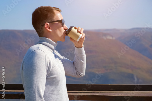 Young guy drinks coffee against mountains on a clear day, sunlight