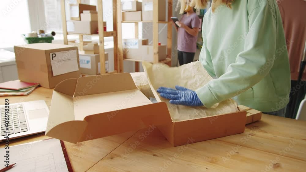 Young blonde woman in protective mask and gloves packing order into box before shipping it to customer while working in office of online store during covid-19 pandemic
