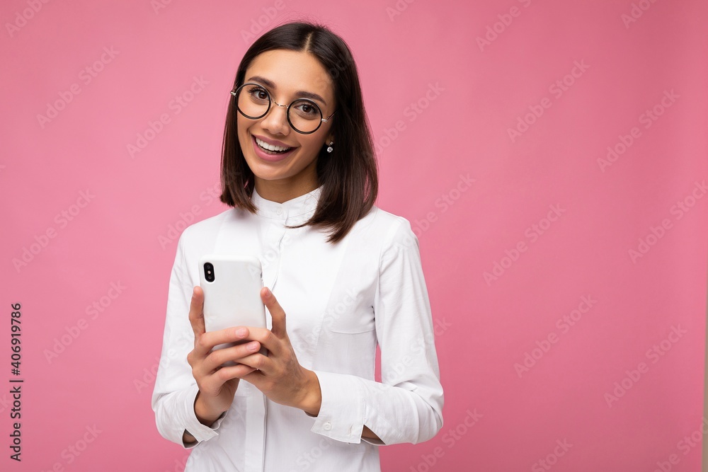Photo of charming smiling young brunette woman wearing white blouse and  optical glasses standing isolated over pink background messaging sms using  mobile phone looking at camera Stock Photo | Adobe Stock