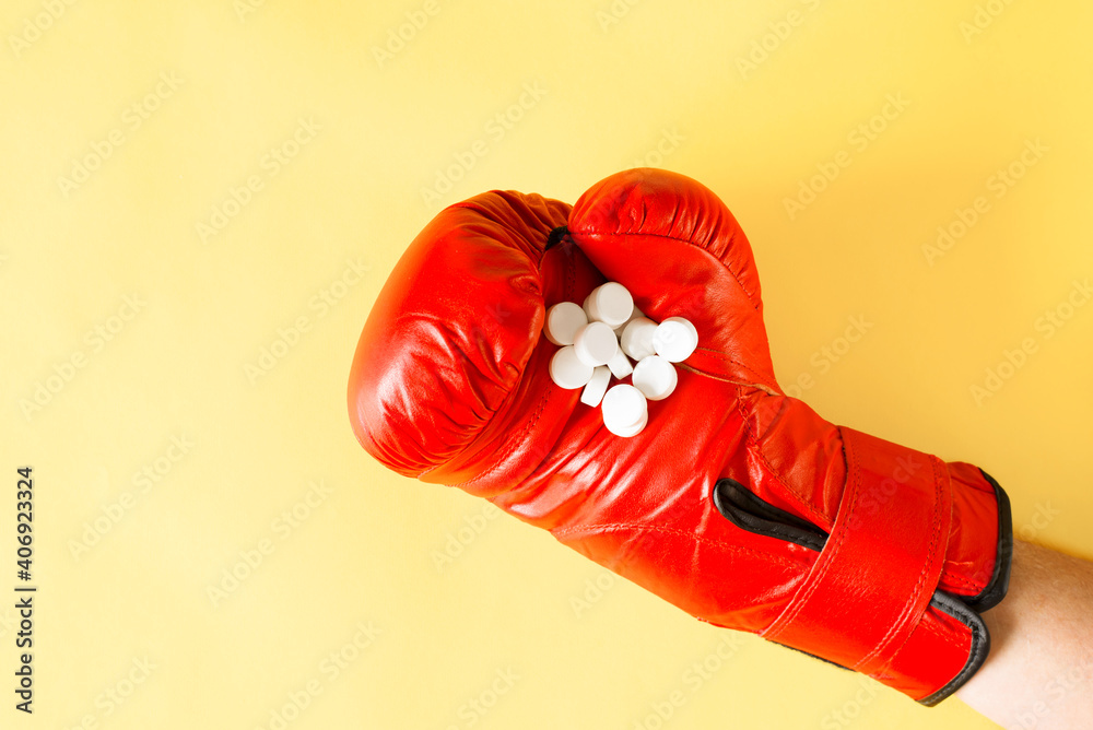 Hand in boxing glove holding pile of pills, close-up. Yellow background, top view.