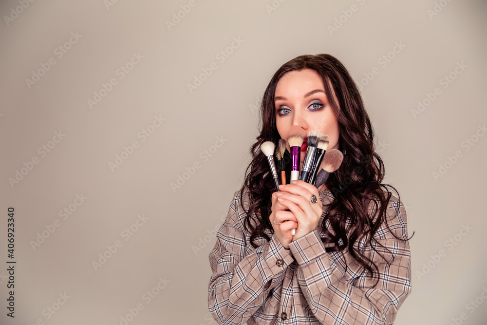Obraz premium Young beautiful woman holding makeup brushes in her hands and posing on grey background at studio.