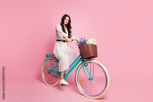 Fototapeta Naklejka Na Ścianę i Meble -  Full length body size photo of female student in white outfit riding bike with flowers smiling happy isolated pastel pink color background