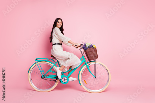 Fototapeta Naklejka Na Ścianę i Meble -  Full length body size photo of pretty woman riding bike with wild flowers in white dress isolated on pastel pink color background