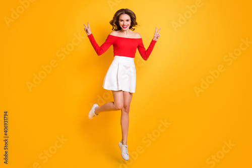 Full length body size photo girl wearing red top white skirt jumping showing v-sign gesture isolated on vivid yellow color background