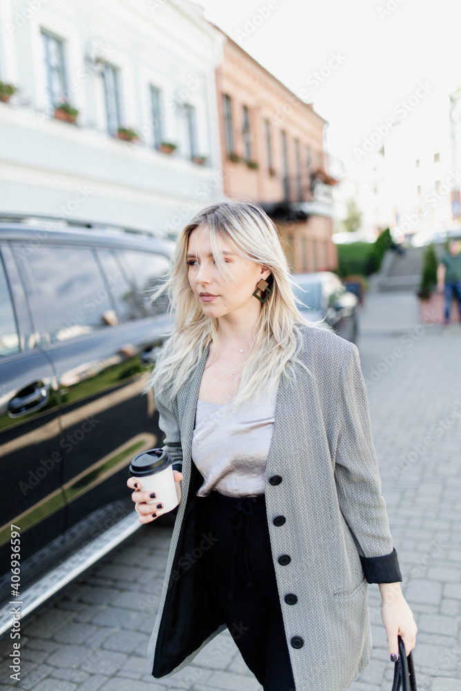 modern young blonde in the city is walking against the background of a car with a cup of coffee.