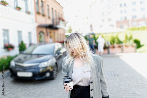 modern young blonde in the city is walking against the background of a car with a cup of coffee.