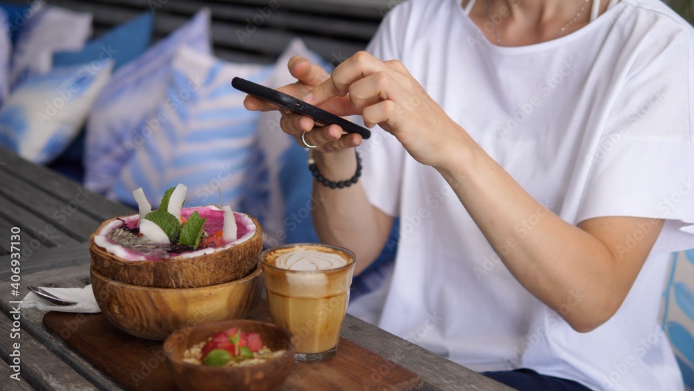 Close up of female hands taking picture of a smoothie served in coconut bowl with granola on a side and coffee. Close up of female hands taking picture of a smoothie served in coconut bowl with granola on a side and coffee.