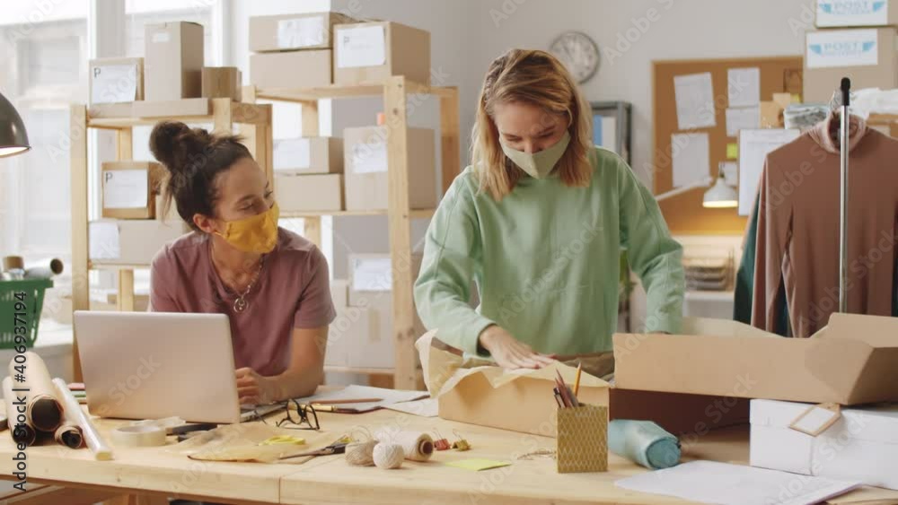 Young woman in protective mask working on laptop in office of online clothes store while her female colleague packing shirt into box before shipping order to customer during coronavirus outbreak