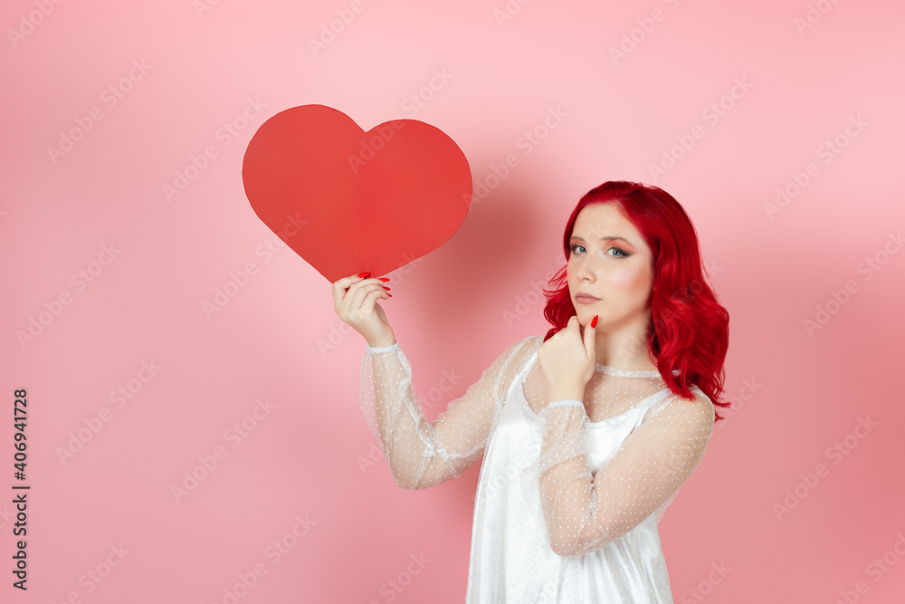 a pensive, doubting, hesitant woman in a white dress and with red hair holds a large paper red heart and rubs her chin with her hand , isolated on a pink background. a pensive, doubting, hesitant woman in a white dress and with red hair holds a large paper red heart and rubs her chin with her hand , isolated on a pink background.