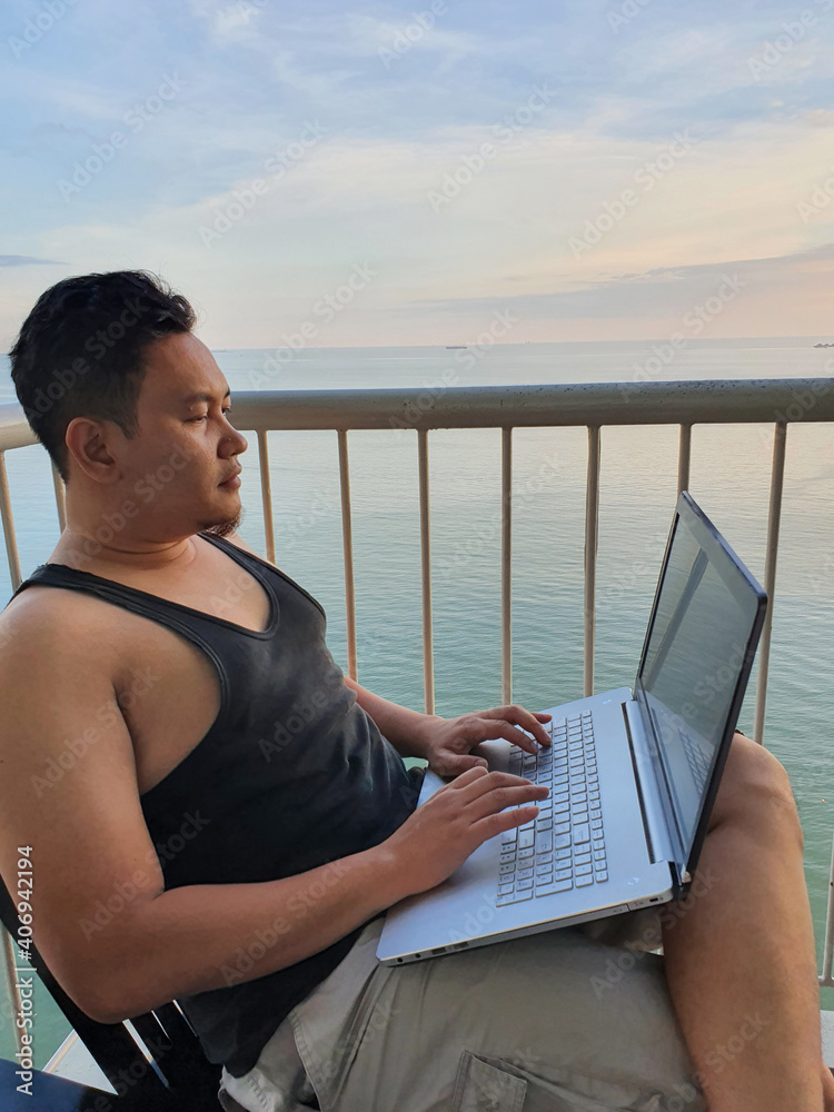 Happy successful Asian man using laptop while sitting on his balcony with calm blue ocean behind, remote working digital nomad