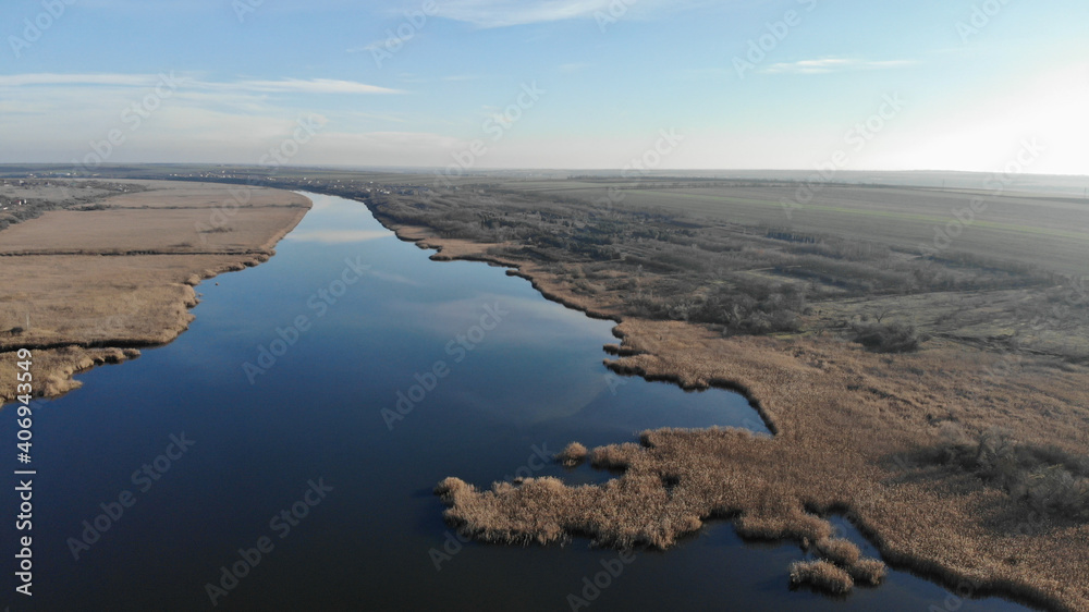 The estuary of a river with blue water in which the sky is reflected. There are dry grass, reeds and trees on the bank of the river. There are village with small houses in the distance