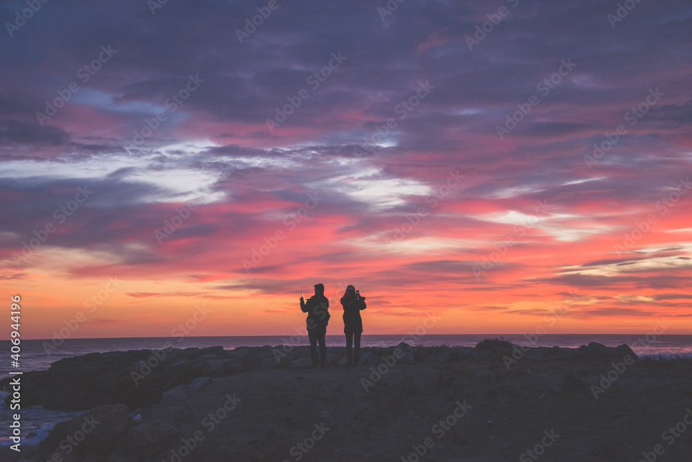 Silueta de pareja haciendo fotografías al atardecer. Nubes rosas y naranjas.