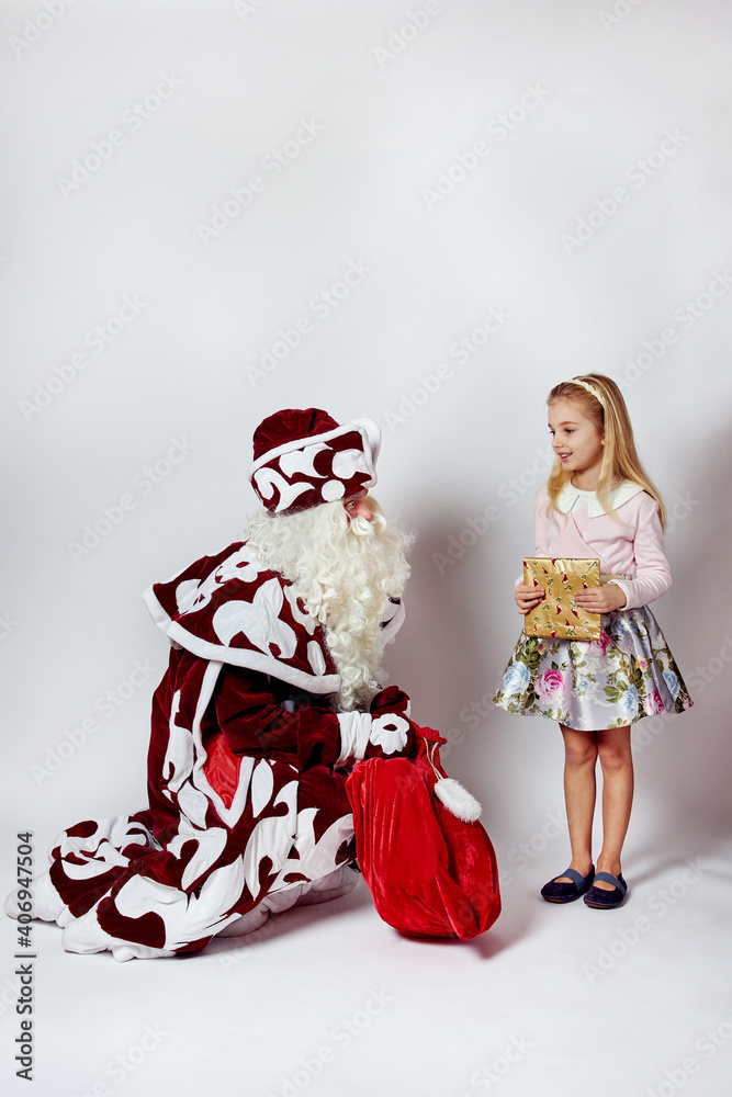 Santa Claus in a red fur coat sits next to him and looks at a cute blonde girl. In the hands of a large bag with gifts. On a white background.