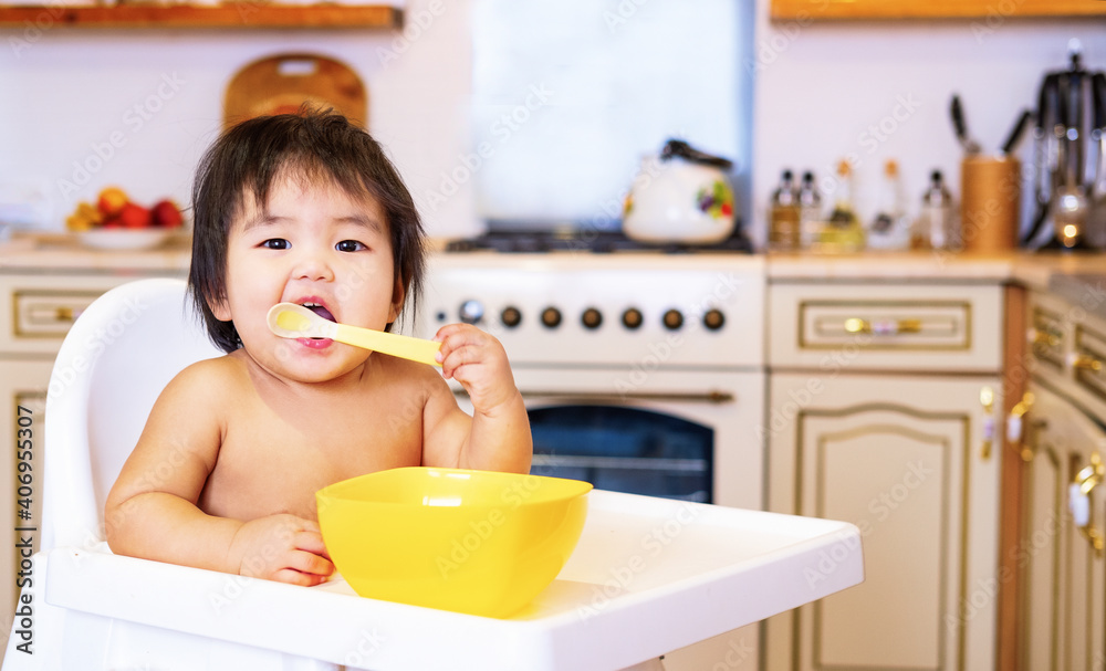 The first feeding of the child. The baby is eating from a yellow spoon. Mom feeds the girl with a spoon. A mother feeds her eight month old baby at home.Children's first meal. Motherhood and childhood The first feeding of the child. The baby is eating from a yellow spoon. Mom feeds the girl with a spoon. A mother feeds her eight month old baby at home.Children's first meal. Motherhood and childhood