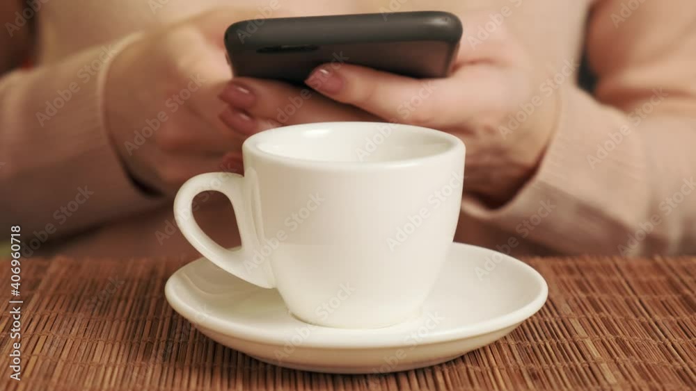 A woman with a mobile phone in her hands and drinking coffee in a white cup. There is a bamboo mat on the table. A woman with a mobile phone in her hands and drinking coffee in a white cup. There is a bamboo mat on the table.