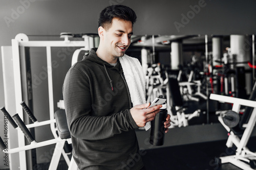Portrait of a handsome man in the gym holding a phone in one hand and smiling, holding a sports water bottle in the other hand