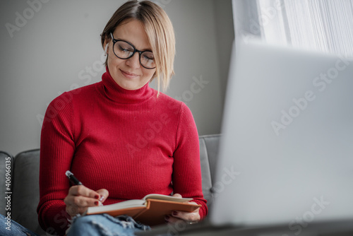 Beautiful woman psychologist wearing glasses at home sitting at home on the couch in front of laptop screen taking notes during video session photo