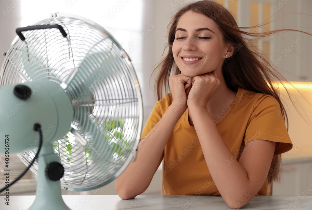 Woman enjoying air flow from fan at table in kitchen. Summer heat - obrazy, fototapety, plakaty 