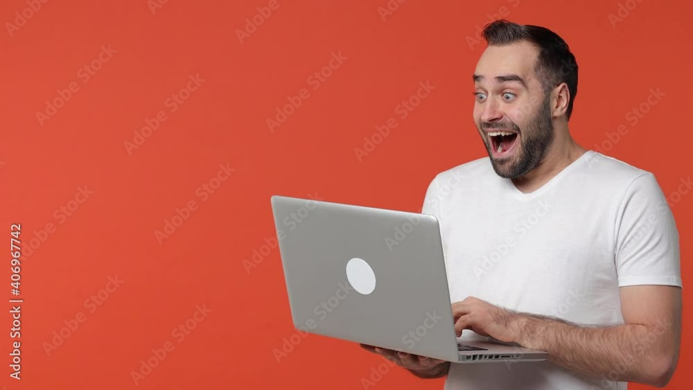 Shocked surprised cheerful bearded young man 20s in white casual t-shirt isolated on orange color background studio. People lifestyle concept. Working on laptop pc computer pointing index finger aside Shocked surprised cheerful bearded young man 20s in white casual t-shirt isolated on orange color background studio. People lifestyle concept. Working on laptop pc computer pointing index finger aside