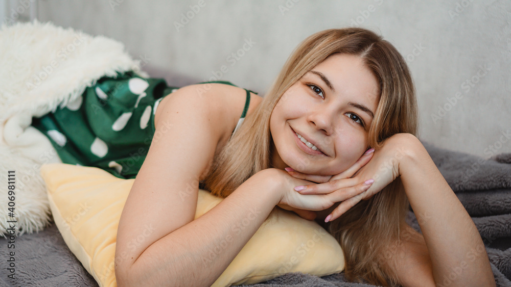 Naklejka premium Young beautiful girl woke up at home. She lies on a yellow pillow and is covered with a white blanket. Blonde smiling