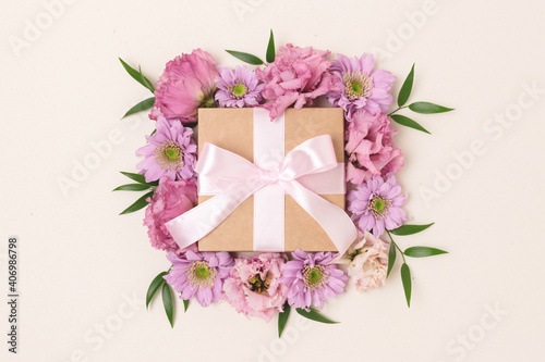 Gift box with a tied ribbon bow and flower frame. Springtime composition on a beige background.