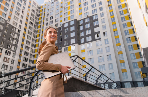 Young successful business woman with a laptop on her hands looks over her shoulder and smiles while climbimg the stairs of a high-rise buiding photo