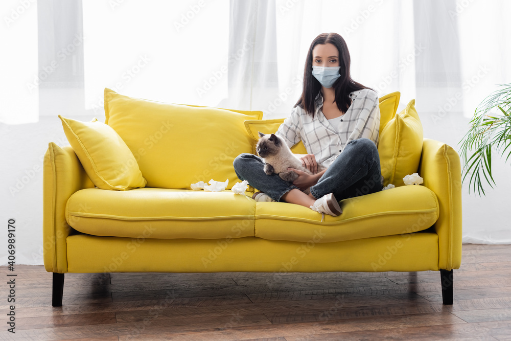 allergic woman in medical mask looking at camera while sitting on sofa with cat near crumpled paper napkins allergic woman in medical mask looking at camera while sitting on sofa with cat near crumpled paper napkins