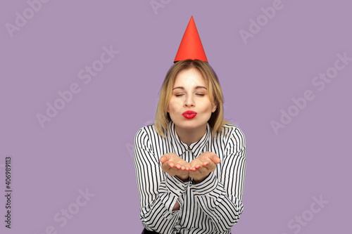 Lovely beautiful woman with funny party cone on head sending sensual air kiss with sexy red lips, sharing love, amorous feelings on Valentines day. indoor studio shot isolated on purple background