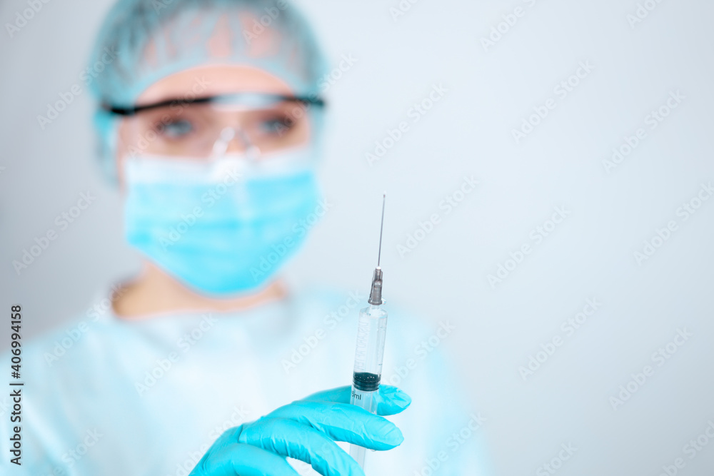 Fototapeta premium A nurse in a medical gown, mask and protective gloves with transparent glasses on her face looks at the needle of a syringe for injection