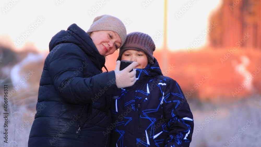 Portrait of a happy mom with her son looking at a photo in a smartphone. Loving family outdoors in winter. People walk in the snowy forest at sunset. Beautiful natural landscape.