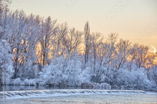 River with frozen banks and trees. Small waterfall in the river with ice. Snowy trees and dramatic sunset