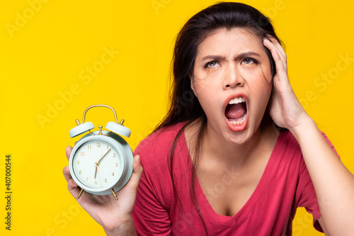Young woman get insomnia She can not sleep until morning Girl holding alarm clock with messy hair Asian lady get unhappy and screaming by open mouth She get frustrated and upset Yellow background