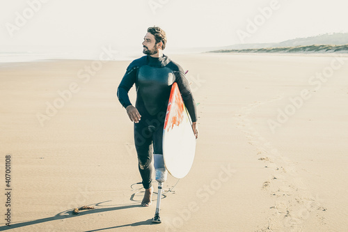 Active surfer with amputated leg walking on beach with surfboard. Bearded amputee in wetsuit pacing on sand, carrying board and looking away. Vertical shot. Artificial limb and extreme sport concept photo