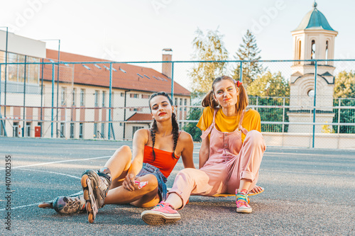 Two teen girls, friends in colorful clothes with roller skates and skate board.