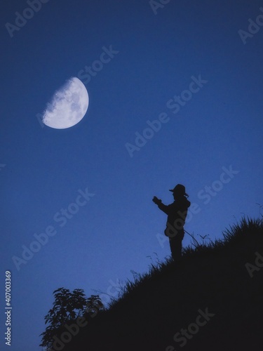 a man on a mountain takes out his cell phone to take a picture of him at sunset while the moon is behind him.