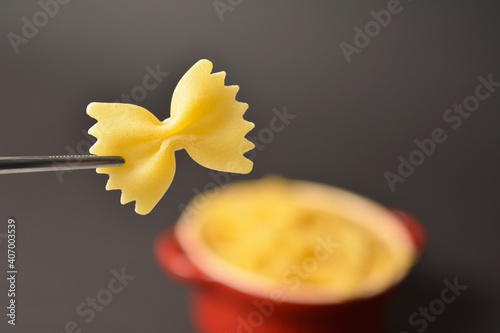 Piece of Uncooked italian farfale bowtie pasta in metal tweezers isolated on black background with red ceramic pan full of raw pasta on background
 photo