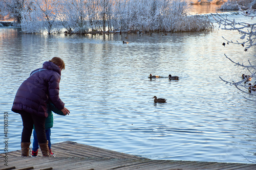 Woman on a wooden pier by the river teaching her son how to feed the ducks.