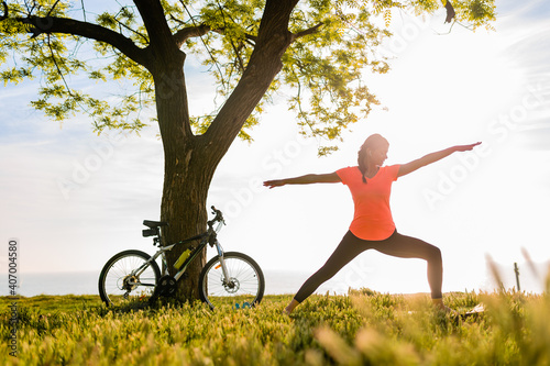 slim beautiful woman silhouette doing sports in morning in park doing yoga on mat in colorful fitness outfit in nature, smiling happy healthy lifestyle, calm meditation, bicycle on background